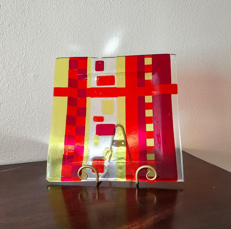May include: Square glass plate with a geometric design in shades of red, yellow, and white. The plate is on a stand.