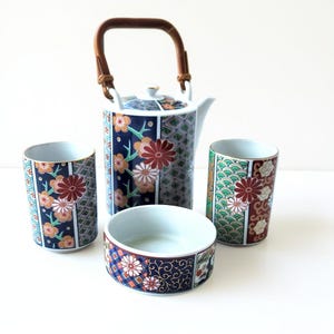 May include: A Japanese-style tea set featuring a teapot with a bamboo handle and two matching cups and a bowl. The set has a navy blue background with floral and geometric patterns in red, white, and gold.