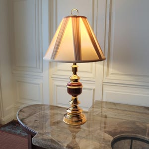 May include: A decorative table lamp with a gold-colored base and a lampshade with vertical beige and brown stripes. The lamp has a dark red accent and sits on a marble-like surface.