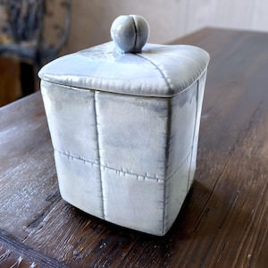 May include: A square ceramic canister with a light blue and grey crackle glaze finish. The container features a grid-like pattern of raised lines resembling stitching and a matching lid topped with a small, rounded, knob-like handle.