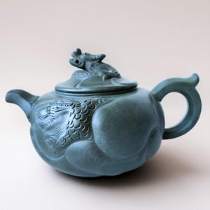 May include: A teal-colored ceramic teapot with a dragon motif. The teapot features a dragon sculpture on the lid and a detailed dragon scale design on the body. The handle is a loop shape. The spout is curved.