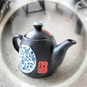 Puede incluir: Pequeña tetera de cerámica negra con tapa y boquilla. La tetera presenta un diseño floral blanco y azul en el lateral y un cuadrado rojo con caracteres chinos. La tapa tiene un pequeño pomo decorativo.
