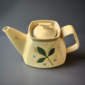 May include: Cream-colored ceramic teapot with a lid, spout, and handle. The teapot features a green leaf design with blue star accents. The teapot is a vintage style and is likely used for brewing tea.