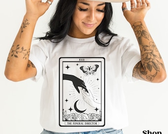 May include: White t-shirt with a black and white tarot card design. The card is labeled "XVIII The Funeral Director" and features a butterfly, a crescent moon, and two hands reaching towards each other.