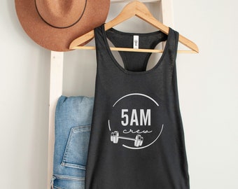 May include: A black tank top with the text "5AM crew" inside a white circle, with a barbell graphic. The tank top is hanging on a wooden hanger. A brown hat and blue jeans are also in the image.