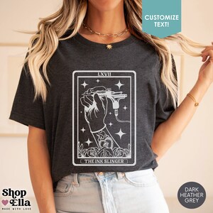 Puede incluir: Camiseta gris oscuro jaspeada con un gr&aacute;fico blanco de una m&aacute;quina de tatuaje y el texto "LXVII THE INK SLINGER".