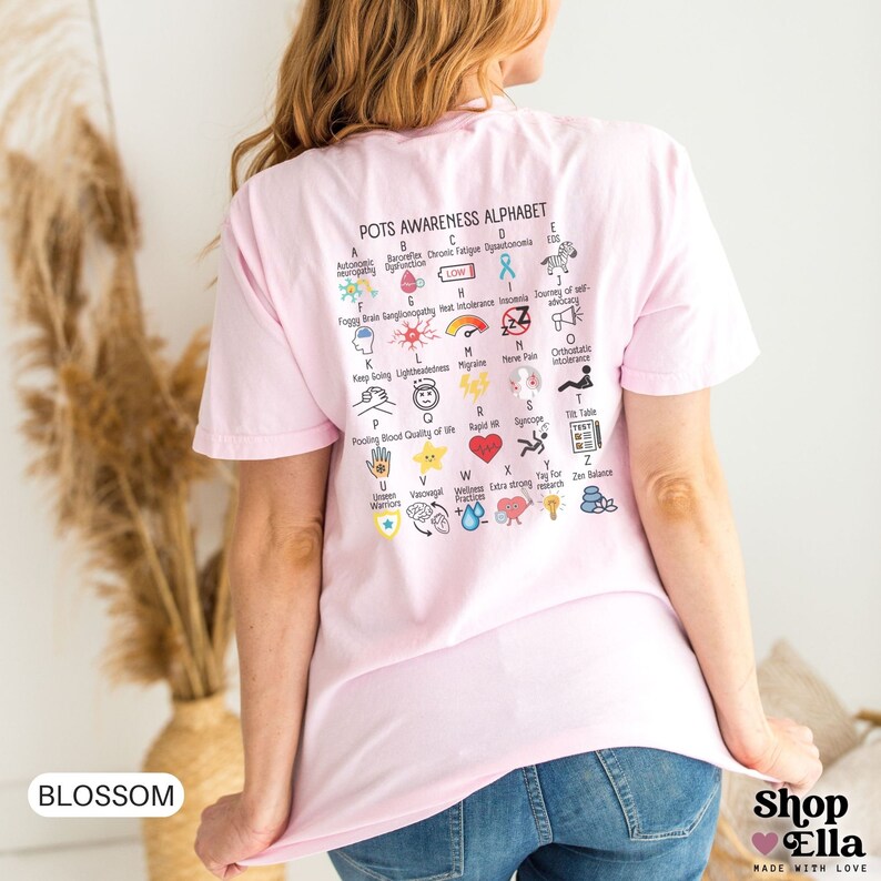May include: A light pink t-shirt with the text "POTS Awareness Alphabet" and various illustrated icons representing different symptoms. The shirt also has the word "BLOSSOM" printed on the bottom left.