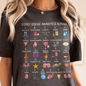 May include: A gray t-shirt with a colorful alphabet chart about kidney disease awareness. Each letter of the alphabet is paired with a term related to kidney health and a corresponding illustration.  The chart includes terms like albuminuria, blood pressure, creatinine, dialysis, edema, filtration, glomerulonephritis, hematuria, immunosuppressant, jaundice, kidney cancer, lupus nephritis, metabolic acidosis, nephrectomy, organ donation, PKD, quality of life, renal failure, stones, transplant, uremia, vasculitis, water balance, extra strong yay research, zest for life.