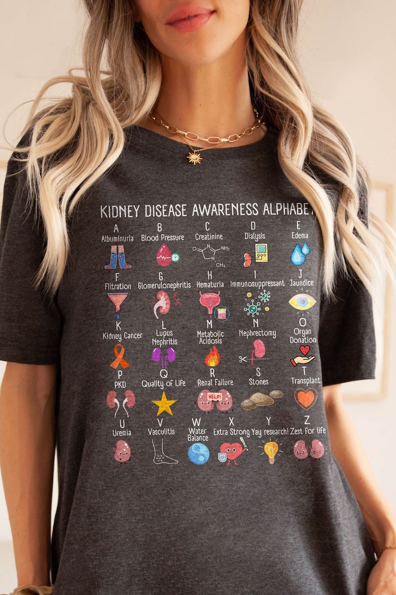 May include: A gray t-shirt with a colorful alphabet chart about kidney disease awareness. Each letter of the alphabet is paired with a term related to kidney health and a corresponding illustration.  The chart includes terms like albuminuria, blood pressure, creatinine, dialysis, edema, filtration, glomerulonephritis, hematuria, immunosuppressant, jaundice, kidney cancer, lupus nephritis, metabolic acidosis, nephrectomy, organ donation, PKD, quality of life, renal failure, stones, transplant, uremia, vasculitis, water balance, extra strong yay research, zest for life.