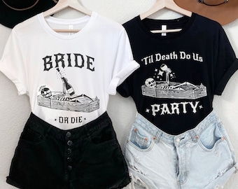 May include: Two white t-shirts with black text and graphics. The first shirt says "Bride" with a skeleton in a coffin holding a bottle of champagne. The second shirt says "'Til Death Do Us Party" with a skeleton in a coffin holding a glass of wine. Both shirts are customizable.