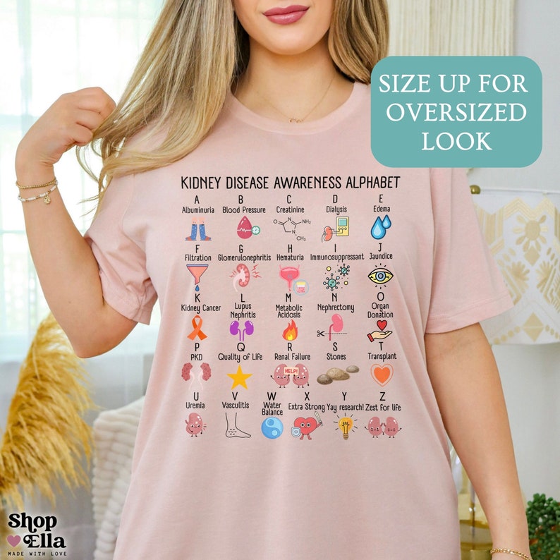 May include: Pink t-shirt with a graphic of a kidney disease awareness alphabet. The alphabet is in white text with colorful icons representing each letter. The text "KIDNEY DISEASE AWARENESS ALPHABET" is at the top of the graphic.