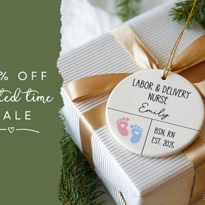K&ouml;nnte beinhalten: Ein runder, wei&szlig;er Ornament mit dem Text "LABOR & DELIVERY NURSE" und "Emily". Es zeigt rosa und blaue Babyf&uuml;&szlig;e und den Text "BSN, RN EST. 2025". Der Ornament h&auml;ngt an einem goldenen Band &uuml;ber einem verpackten Geschenk.