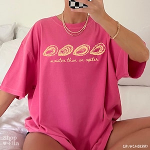 May include: A bright pink t-shirt with the text "moister than an oyster" in a yellow font. Above the text are four oyster shell illustrations. The shirt is a short-sleeved, oversized style.