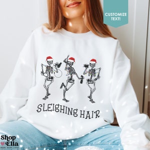 May include: White sweatshirt with a Christmas-themed design featuring three skeletons wearing Santa hats and holding hairdressing tools. The text "SLEIGHING HAIR" is printed below the skeletons.