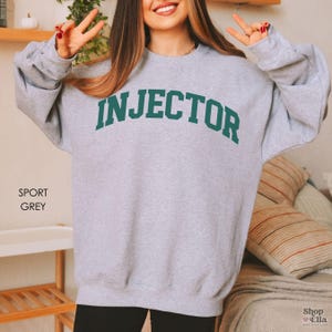 May include: A gray sweatshirt with the word "INJECTOR" printed in green on the front. The sweatshirt is a relaxed fit and has a crew neck.