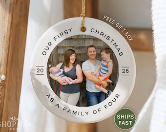 May include: A white ceramic Christmas ornament with a family photo inside. The ornament has the text "Our First Christmas as a Family of Four" and the years "20" and "26". A gold string is attached for hanging.