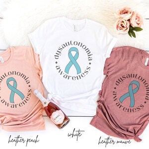 May include: Three t-shirts in different colours, heather peach, white, and heather mauve, with a teal ribbon and the text "dysautonomia awareness" printed on each.