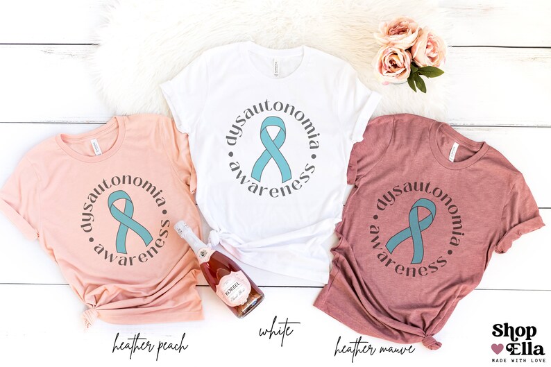 May include: Three t-shirts in different colours, heather peach, white, and heather mauve, with a teal ribbon and the text "dysautonomia awareness" printed on each.