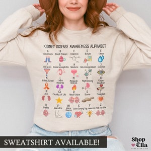 May include: A beige sweatshirt with a graphic print of an alphabet representing kidney disease awareness. The alphabet includes terms like albuminuria, blood pressure, creatinine, dialysis, edema, filtration, glomerulonephritis, hematuria, immunosuppressant, jaundice, kidney cancer, lupus nephritis, metabolic acidosis, nephrectomy, organ donation, PKD, quality of life, renal failure, stones, transplant, uremia, vasculitis, water balance, extra strong yay research, and zest for life. The sweatshirt has the text "SWEATSHIRT AVAILABLE!" printed at the bottom.