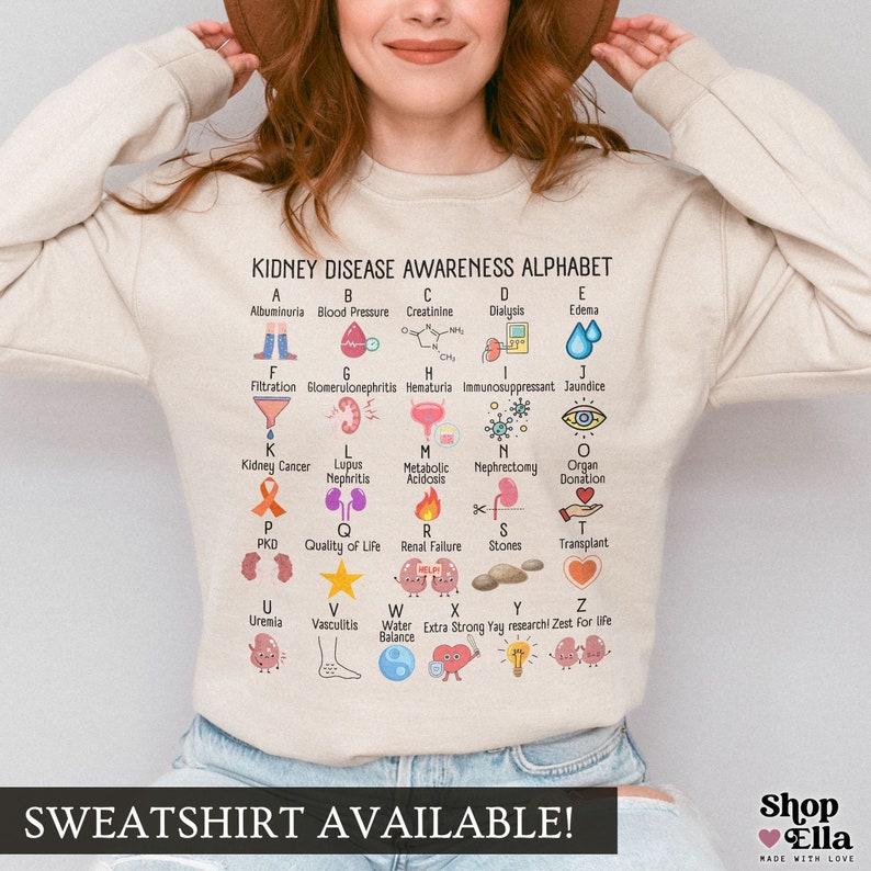 May include: A beige sweatshirt with a graphic print of an alphabet representing kidney disease awareness. The alphabet includes terms like albuminuria, blood pressure, creatinine, dialysis, edema, filtration, glomerulonephritis, hematuria, immunosuppressant, jaundice, kidney cancer, lupus nephritis, metabolic acidosis, nephrectomy, organ donation, PKD, quality of life, renal failure, stones, transplant, uremia, vasculitis, water balance, extra strong yay research, and zest for life. The sweatshirt has the text "SWEATSHIRT AVAILABLE!" printed at the bottom.