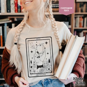 May include: A beige t-shirt with a black and white illustration of a hand holding an apple. The illustration is in the style of a tarot card and includes the text "XCII" and "ELL TEACHER".