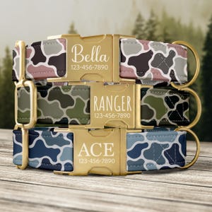 Three dog collars with camouflage patterns in pink, green, and blue. Each collar features a gold-tone metal buckle engraved with a name and phone number: ZOE 123-456-7890, RANGER 123-456-7890, and Milo 123-456-7890. Each includes a gold D-ring.