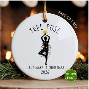 May include: A round white ceramic ornament featuring a black silhouette of a person in a yoga tree pose. The figure is wrapped in colorful Christmas lights and holds a yellow lotus flower. Text reads TREE POSE ...BUT MAKE IT CHRISTMAS 2026.