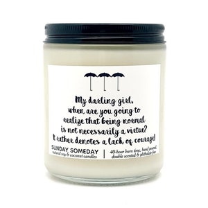 May include: A white candle in a glass jar with a black lid. The label features the text "My darling girl, when are you going to realize that being normal is not necessarily a virtue?" and three umbrella icons.