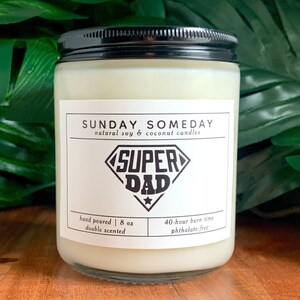 May include: A clear glass jar candle with a black lid. The label reads "SUNDAY SOMEDAY" and "SUPER DAD". The candle is hand-poured, double scented, and contains 8 oz. It has a 40-hour burn time and is phthalate-free.