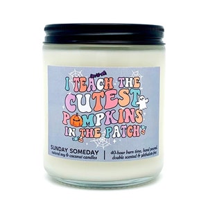 Puede incluir: Una vela en frasco de vidrio transparente con tapa negra, rellena de cera blanca. La etiqueta muestra el texto I TEACH THE CUTEST PUMPKINS IN THE PATCH en letras retro coloridas, rodeado de ilustraciones de una calabaza, fantasma y telarañas.