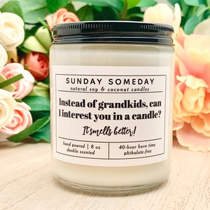 May include: A clear glass jar candle with a black lid. The label reads "SUNDAY SOMEDAY" and "Instead of grandkids, can I interest you in a candle? It smells better!" The candle is 8 oz and has a 40-hour burn time. The background features flowers.