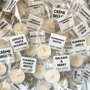 Puede incluir: Un surtido de pequeñas velas redondas de color crema en envases de plástico transparente. Cada vela tiene una etiqueta blanca con un nombre de aroma como "Pumpkin Apple" o "Marshmallow Fireside". Las etiquetas también incluyen el texto "Sunday Someday".