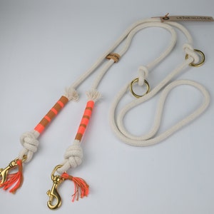 May include: White cotton rope dog lead with two orange and pink striped handles and gold metal clips. The lead has a brown leather tag with the text "LEINENLUDER" on it.