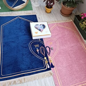 May include: Two prayer rugs, one blue and one pink, with gold trim and tassels. A white gift box with a heart-shaped cutout and the letter "M" embroidered on the blue rug.