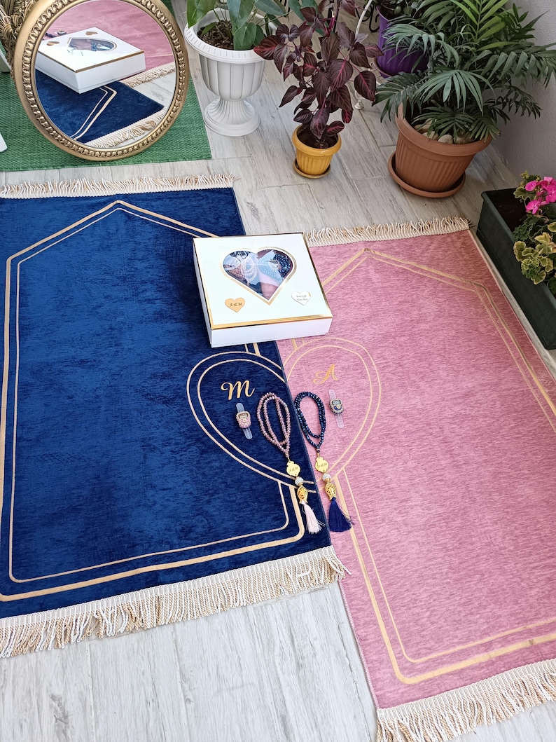 May include: Two prayer rugs, one blue and one pink, with gold trim and tassels. A white gift box with a heart-shaped cutout and the letter "M" embroidered on the blue rug.