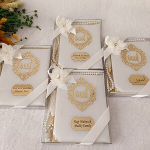 Pu&ograve; includere: Quattro scatole regalo rettangolari bianche contenenti libri di preghiera, ciascuna decorata con un emblema di calligrafia araba dorata, un fiore di stoffa bianco, un nastro bianco e un rosario di perle bianche. Ogni scatola ha una targhetta dorata.