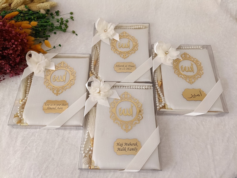 Pu&ograve; includere: Quattro scatole regalo rettangolari bianche contenenti libri di preghiera, ciascuna decorata con un emblema di calligrafia araba dorata, un fiore di stoffa bianco, un nastro bianco e un rosario di perle bianche. Ogni scatola ha una targhetta dorata.