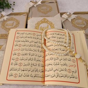 K&ouml;nnte beinhalten: Ein aufgeschlagener Koran mit cremefarbenen Seiten, schwarzem arabischem Text und roten Akzenten, drapiert mit einer Kette aus wei&szlig;en Perlen-Gebetsketten. Im Hintergrund sind mehrere Geschenkboxen mit goldenem Emblem und wei&szlig;er Schleife zu sehen.