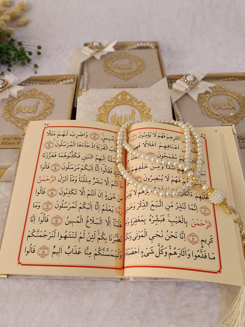 K&ouml;nnte beinhalten: Ein aufgeschlagener Koran mit cremefarbenen Seiten, schwarzem arabischem Text und roten Akzenten, drapiert mit einer Kette aus wei&szlig;en Perlen-Gebetsketten. Im Hintergrund sind mehrere Geschenkboxen mit goldenem Emblem und wei&szlig;er Schleife zu sehen.