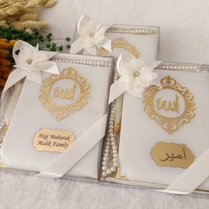 Pu&ograve; includere: Tre scatole regalo rettangolari bianche contenenti grani da preghiera e un libro, ciascuna decorata con un fiore in tessuto bianco, un emblema in filigrana dorata con calligrafia araba e una targhetta dorata con testo personalizzato.