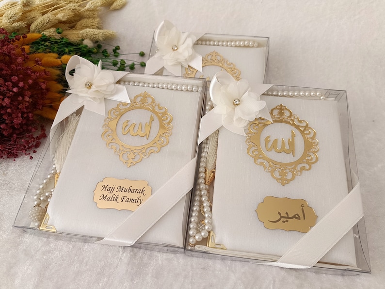 Pu&ograve; includere: Tre scatole regalo rettangolari bianche contenenti grani da preghiera e un libro, ciascuna decorata con un fiore in tessuto bianco, un emblema in filigrana dorata con calligrafia araba e una targhetta dorata con testo personalizzato.