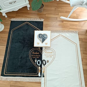 May include: Two black and white prayer rugs with gold trim and tassels. The rugs are personalized with the names "Zubair" and "Zainab" and a quote from the Quran. The rugs are accompanied by a white box with a heart-shaped opening and a gold trim. The box contains a pair of prayer beads, a watch, and a small decorative item.