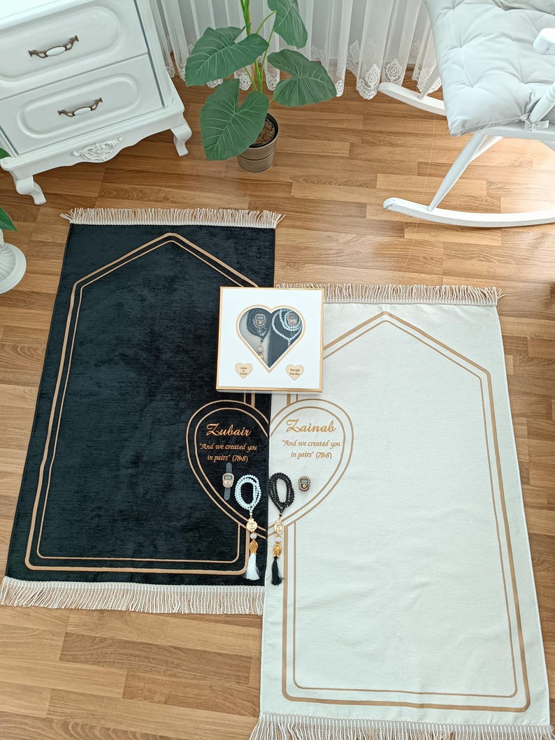 May include: Two black and white prayer rugs with gold trim and tassels. The rugs are personalized with the names "Zubair" and "Zainab" and a quote from the Quran. The rugs are accompanied by a white box with a heart-shaped opening and a gold trim. The box contains a pair of prayer beads, a watch, and a small decorative item.