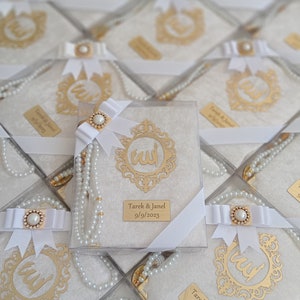 Puede incluir: Cajas de regalo blancas con tapas transparentes, cada una contiene una tela blanca con un diseño de caligrafía árabe dorada, una cadena de perlas blancas y una cinta blanca con un adorno de perla. Las cajas tienen una etiqueta blanca con el texto "Tarek & Janel 9/9/2023".