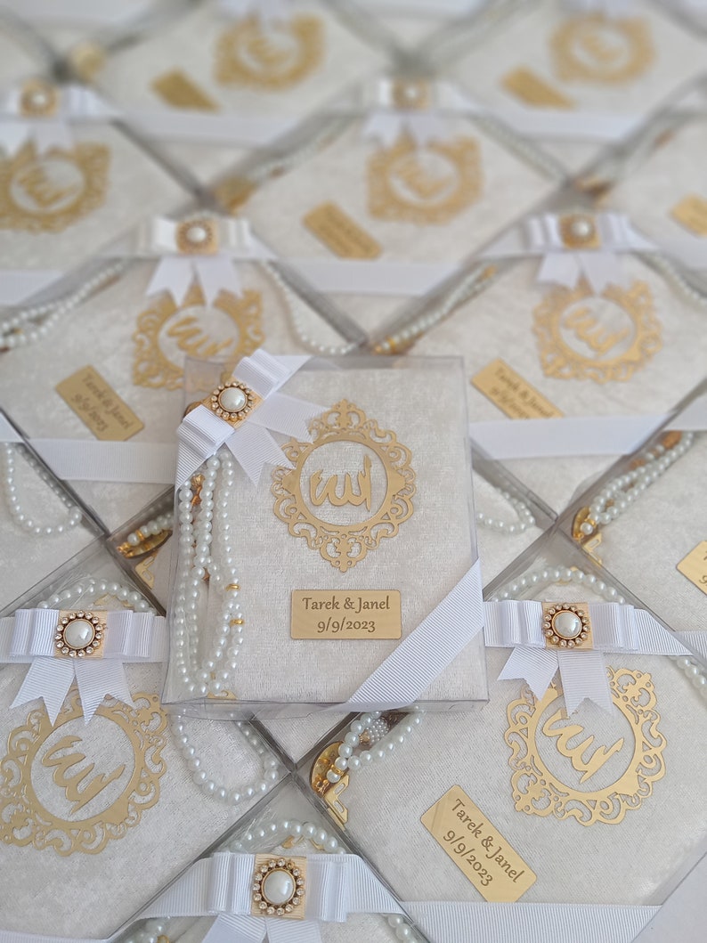 K&ouml;nnte beinhalten: Eine quadratische Geschenkbox mit einem Gebetbuch mit wei&szlig;em, strukturiertem Einband, einem goldenen, verzierten Rahmen mit arabischer Kalligrafie und einem wei&szlig;en Band mit Perlenverzierung. Eine goldene Plakette zeigt Tarek & Janel 9/9/2023.