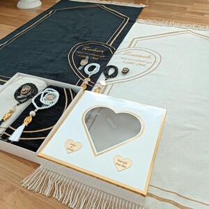May include: Two white prayer rugs with gold trim and tassels. The rugs have the names "Zubair" and "Zainab" embroidered on them, along with the text "And we created you in pairs" (2008). The rugs are accompanied by a white gift box with a heart-shaped window and a gold heart with the names "Zubair" and "Zainab" on it. The text "Your Gift Note Here" is also on the box.