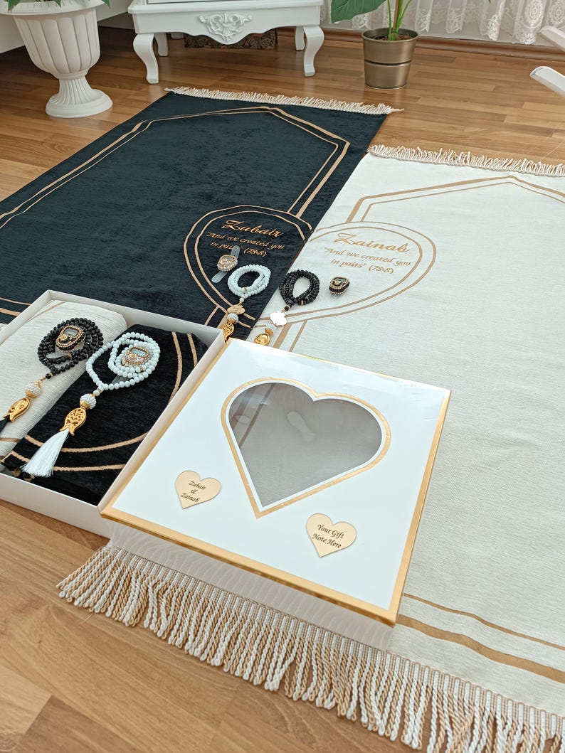 May include: Two white prayer rugs with gold trim and tassels. The rugs have the names "Zubair" and "Zainab" embroidered on them, along with the text "And we created you in pairs" (2008). The rugs are accompanied by a white gift box with a heart-shaped window and a gold heart with the names "Zubair" and "Zainab" on it. The text "Your Gift Note Here" is also on the box.