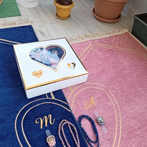 May include: Two prayer rugs, one blue and one pink, with gold trim and embroidered letters. Each rug has a matching prayer bead set and a digital tasbih counter. The rugs are packaged in a white gift box with a heart-shaped window.