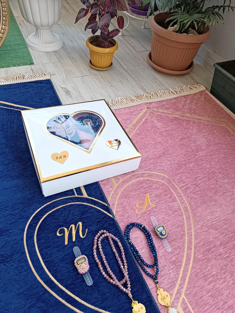 May include: Two prayer rugs, one blue and one pink, with gold trim and embroidered letters. Each rug has a matching prayer bead set and a digital tasbih counter. The rugs are packaged in a white gift box with a heart-shaped window.