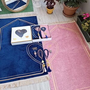 May include: Two prayer rugs, one blue and one pink, with gold trim and tassels. Each rug has a heart-shaped design with a gold outline. A small white box with a heart-shaped cutout is in the center of the image. The box contains two sets of prayer beads.